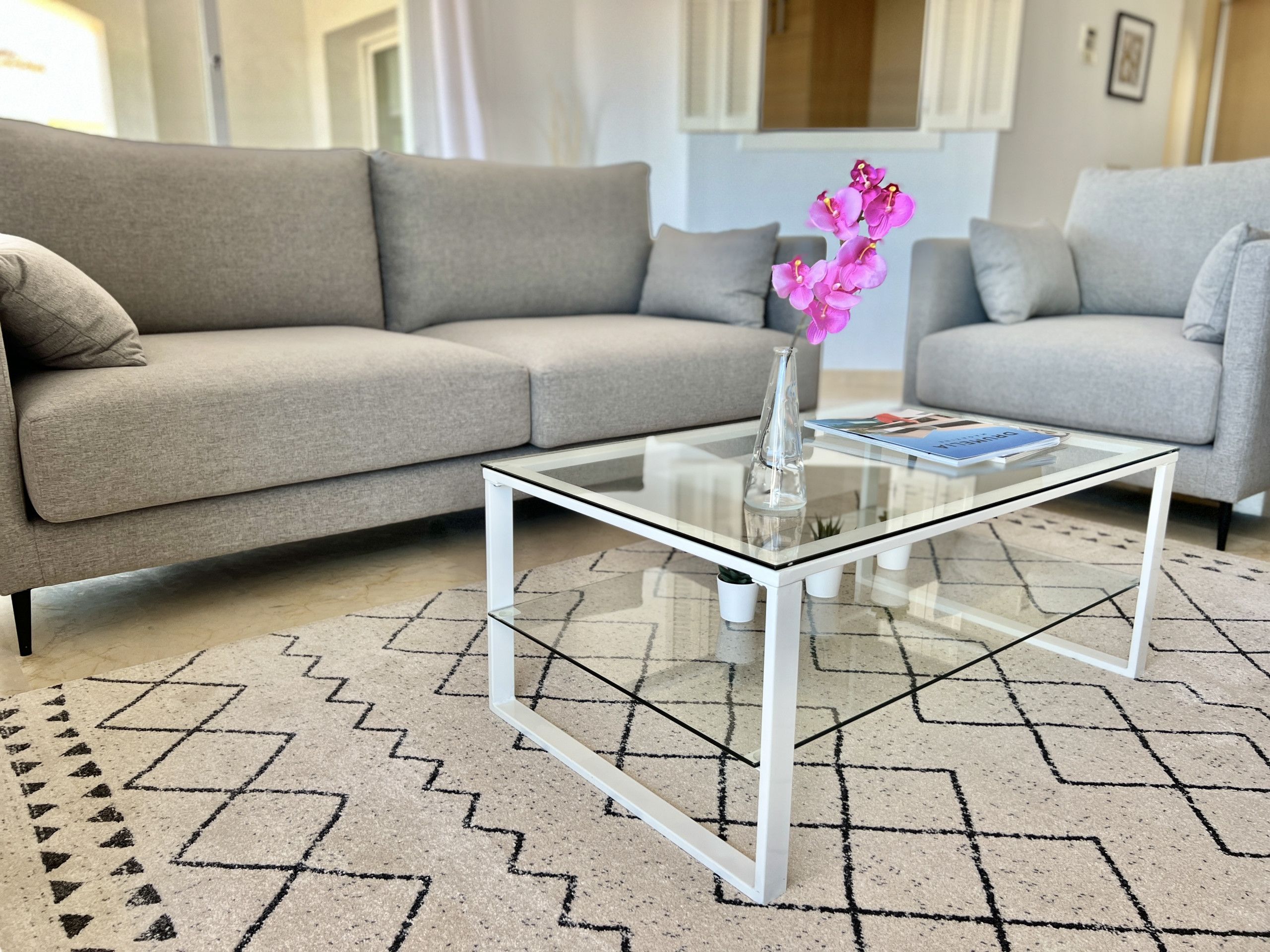 Een woonkamer met een glazen salontafel, een beige bank en een kamerbreed tapijt met een patroon.