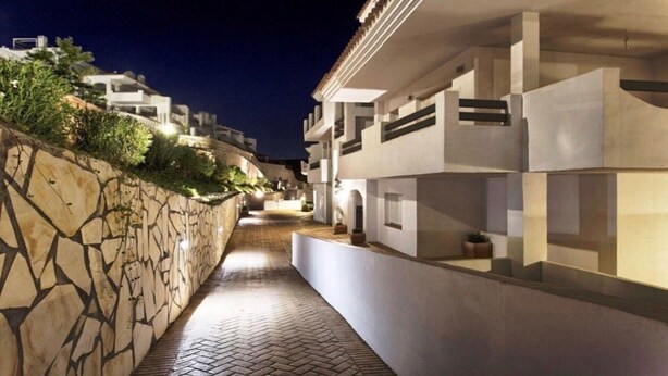 Uma passagem estreita ladeada por edifícios modernos com paredes de pedra e plantas em vasos.