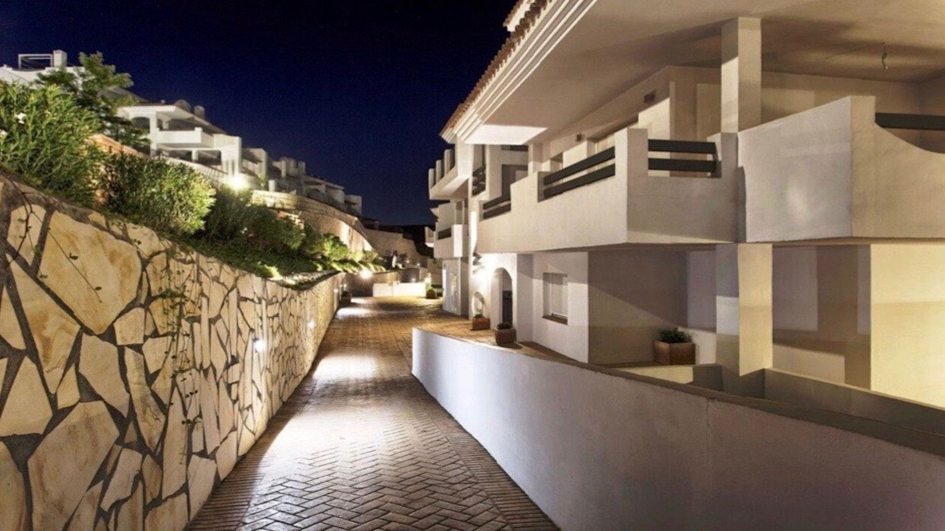 Un sendero estrecho bordeado por edificios modernos con muros de piedra y plantas en macetas.