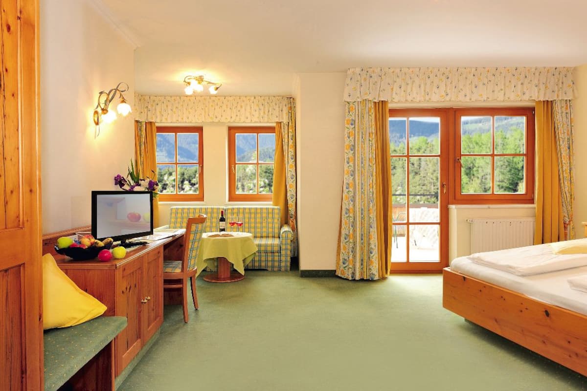 Ein gemütliches Schlafzimmer mit einem hölzernen Bett, einem kleinen Tisch mit Stuhl, einem Fernseher und einem Fenster mit Vorhängen.