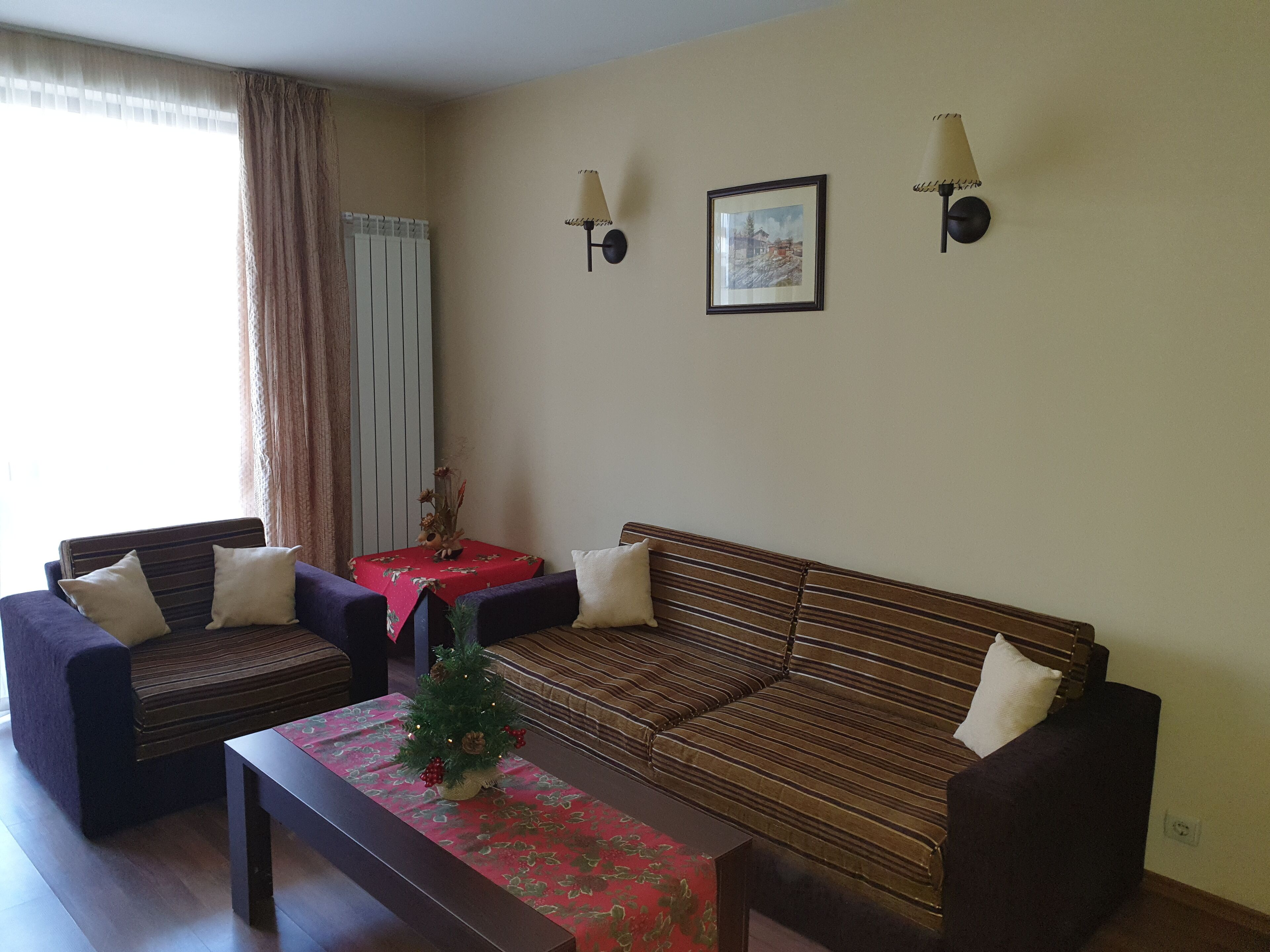 A living room with a sofa, two armchairs, a coffee table, and a small table with a vase. There are two wall-mounted lamps and a framed picture on the wall.