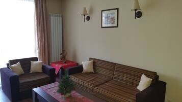 A living room with a sofa, two armchairs, a coffee table, and a small table with a vase. There are two wall-mounted lamps and a framed picture on the wall.