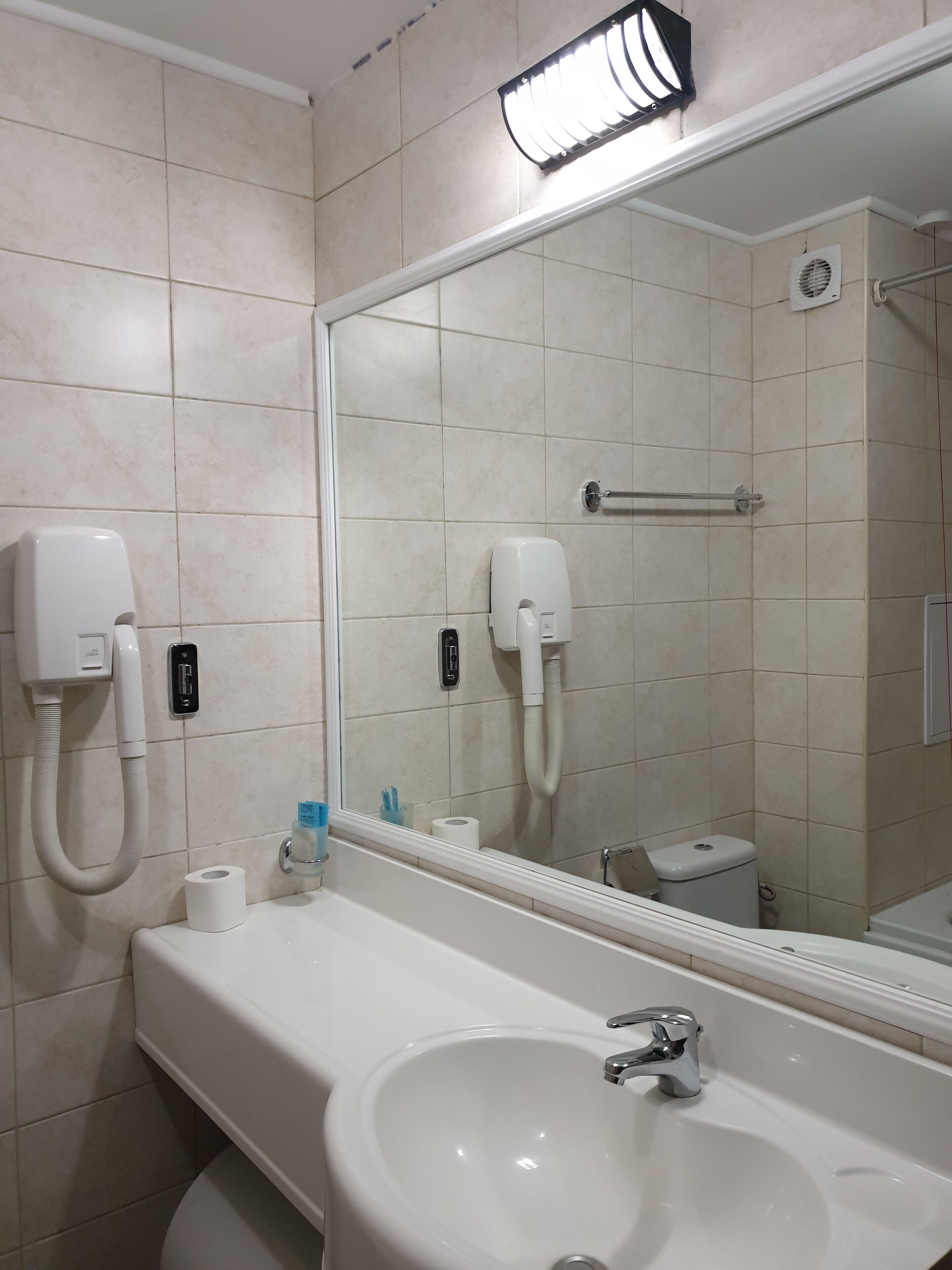 Un baño con lavabo, espejo, toallero y dos secadores de manos.