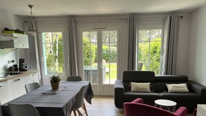 A modern living room with a dining area, a black sofa with 'Charme' pillows, a red chair, and a view of greenery through large windows.
