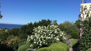 Un jardín con diversas plantas y flores, un césped bien cuidado y vista al mar de fondo.