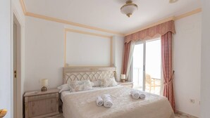 A bedroom with a large bed, a nightstand, a lamp, and a view of the outdoors. - Apartamaneto Joya 4 by Costa CarpeDiem (Calp)