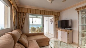 A modern living room with a large beige sectional sofa, a glass coffee table, a flat-screen TV on a decorative cabinet, and a view of the outdoors through large windows. - Apartamaneto Joya 4 by Costa CarpeDiem (Calp)