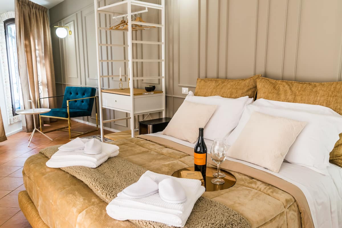 A neatly made bed with a bottle of wine and a glass, a blue armchair, and a white wardrobe in a well-lit room with a view of the outdoors.