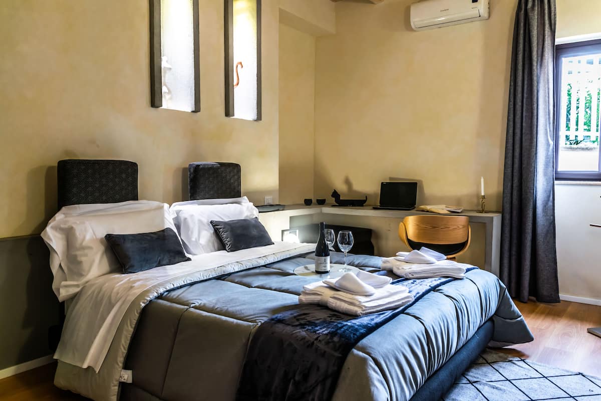 A neatly made bed with a blue and white duvet, a bedside table with a bottle and glasses, a chair, and a desk with a computer.