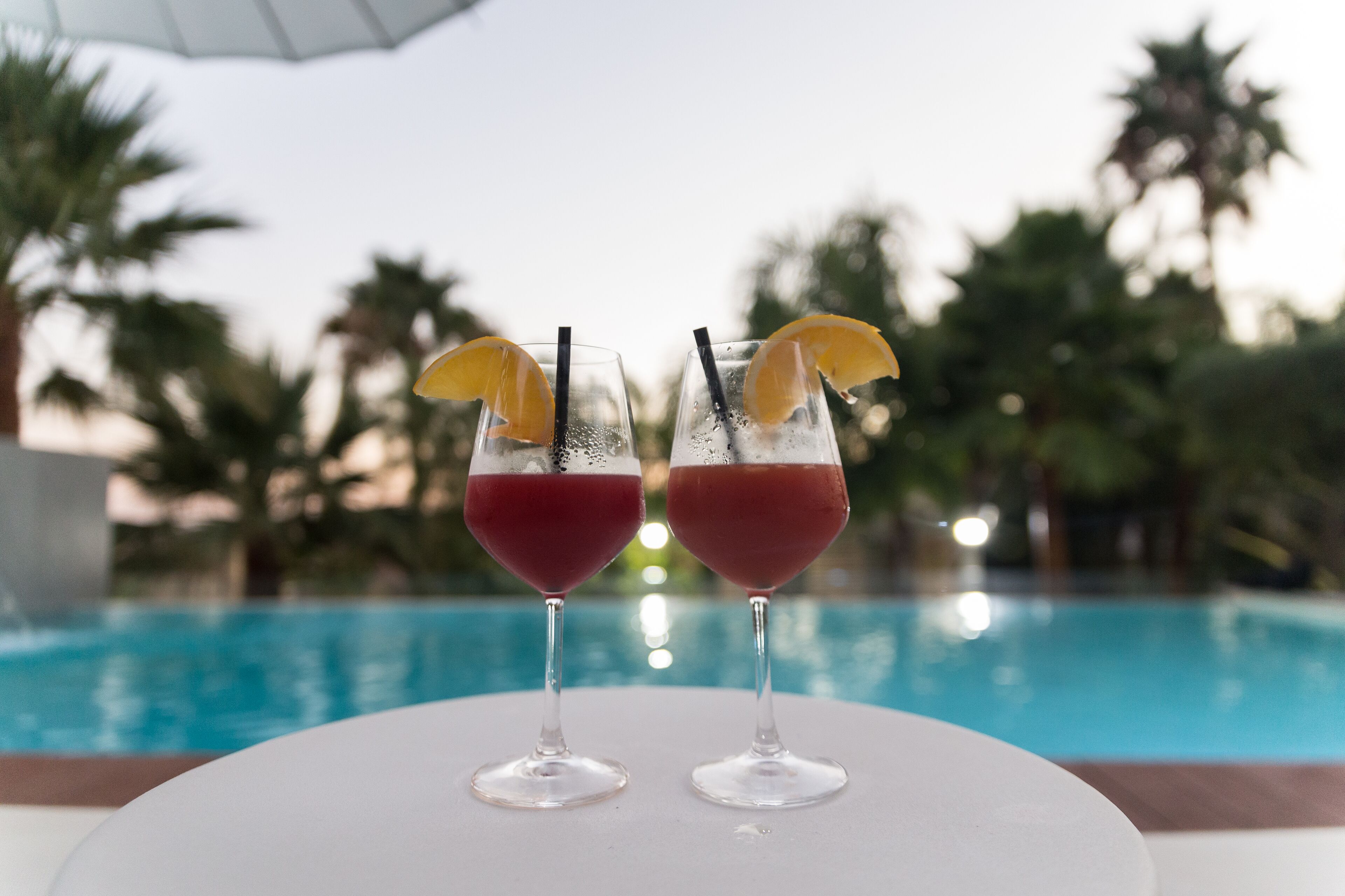 Dos vasos de bebida roja con rodajas de limón y pajitas sobre una mesa blanca junto a una piscina con palmeras al fondo.