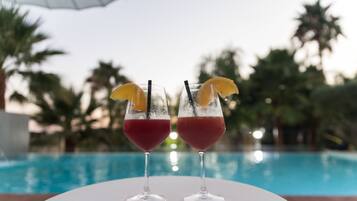 Dos vasos de bebida roja con rodajas de limón y pajitas sobre una mesa blanca junto a una piscina con palmeras al fondo.