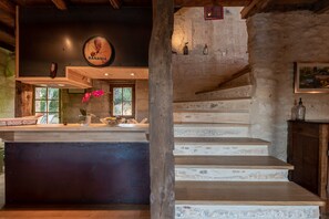Un interior rústico con una escalera de madera, una zona de cocina con un reloj y una ventana que da a zonas verdes.