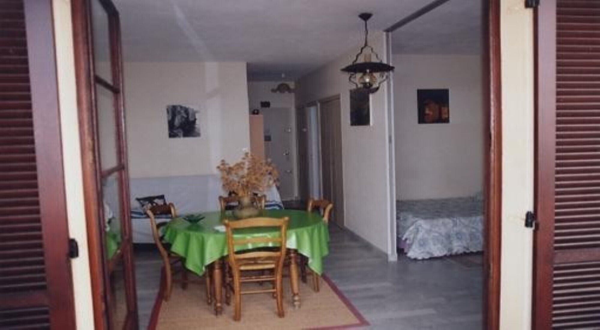 Un coin repas avec une table dressée pour quatre personnes, une nappe verte et un vase de fleurs. On aperçoit, au fond, un lit recouvert d’une courtepointe blanche.