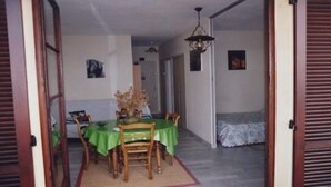 Un coin repas avec une table dressée pour quatre personnes, une nappe verte et un vase de fleurs. On aperçoit, au fond, un lit recouvert d’une courtepointe blanche.