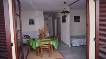 Un coin repas avec une table dressée pour quatre personnes, une nappe verte et un vase de fleurs. On aperçoit, au fond, un lit recouvert d’une courtepointe blanche.