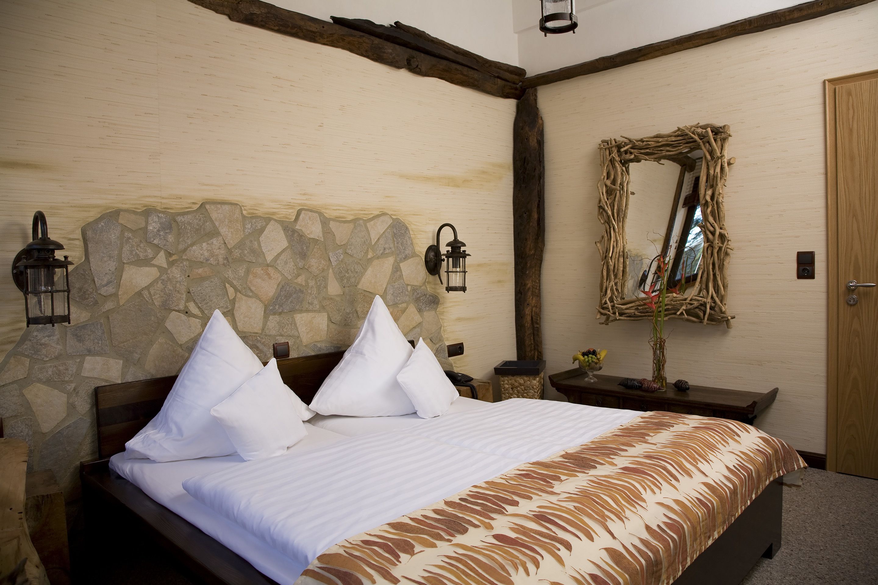 Un lit bien fait, avec du linge de lit blanc et une couvre-lit à motifs, un mur en pierre avec une poutre en bois au plafond et un miroir rustique accroché au mur.