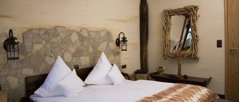 A neatly made bed with white linens and a patterned bedspread, a stone wall with a wooden beam ceiling, and a rustic mirror on the wall.