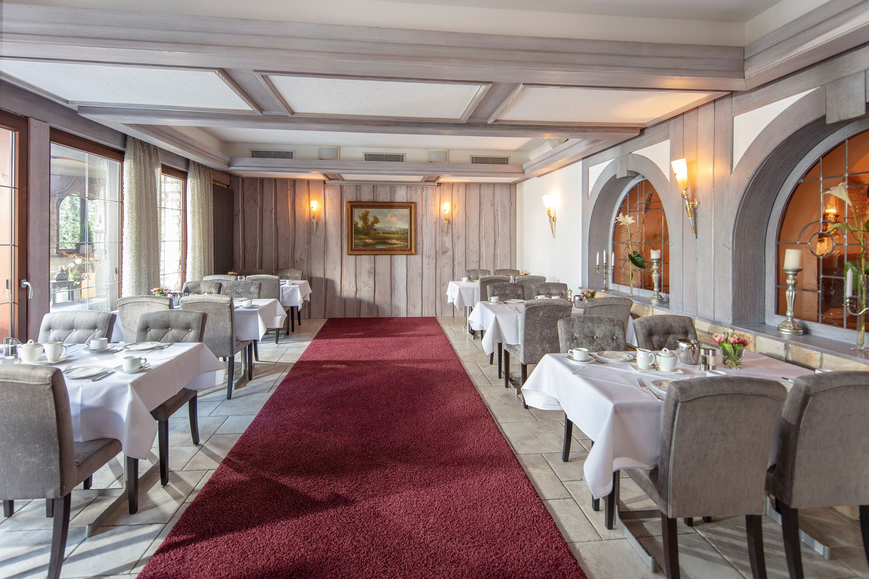 A formal dining room with tables set for service, a red carpet, and a painting on the wall.