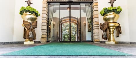 Ein Hotelzugang mit einem steinernen Bogen, einem grünen Teppich und zwei großen Goldvasen mit Pflanzen und Schleifen.