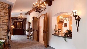 A grand hallway with a chandelier, wooden doors, a stone wall, and a reception area with a clock and decorative items.