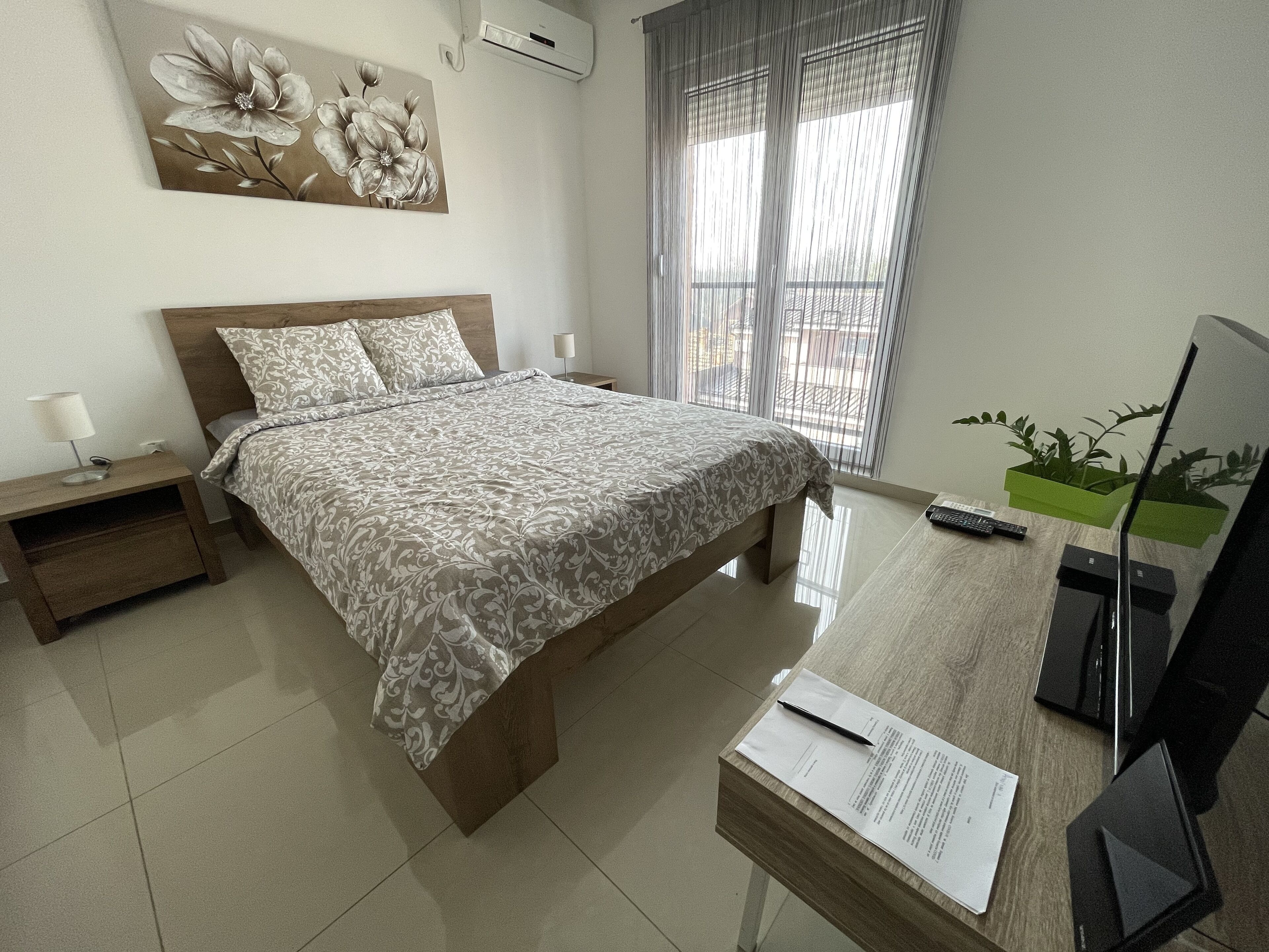 A neatly arranged bedroom with a bed, bedside tables, a desk with a computer, and a window with curtains.