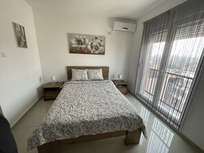 A bedroom with a bed, nightstands, a wall-mounted air conditioner, and a window with blinds. - King Peter Apartments (Sremska Mitrovica)