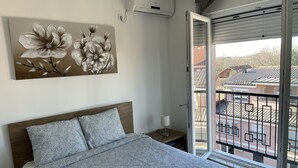 A bedroom with a bed, a nightstand, a lamp, a wall-mounted air conditioner, and a large window with a view of buildings.