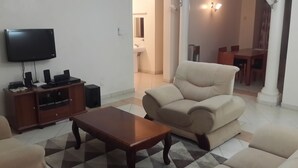 A living room with a flat-screen TV, a wooden coffee table, a beige sofa, and a dining area in the background. - Shikara Apartment (Mombasa)