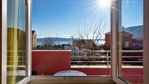 A balcony with a view of the sea and buildings, a red wall, and a metal railing.