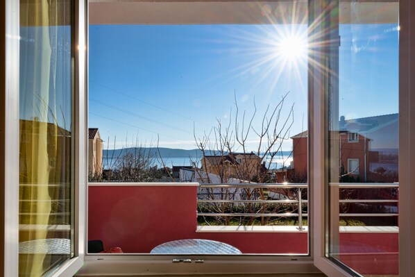 A balcony with a view of the sea and buildings, a red wall, and a metal railing.