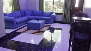 Een woonkamer met een blauwe bank, een klein voetenbankje, een eettafel met stoelen en een uitzicht op gebouwen door grote ramen.