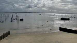 A beach with a wooden walkway leading to the water, boats in the distance, and a cloudy sky. - House / Villa - Cap ferret (Lège-Cap-Ferret)