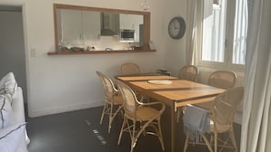 A dining area with a wooden table and wicker chairs, a kitchen in the background, and a clock on the wall. - House / Villa - Cap ferret (Lège-Cap-Ferret)