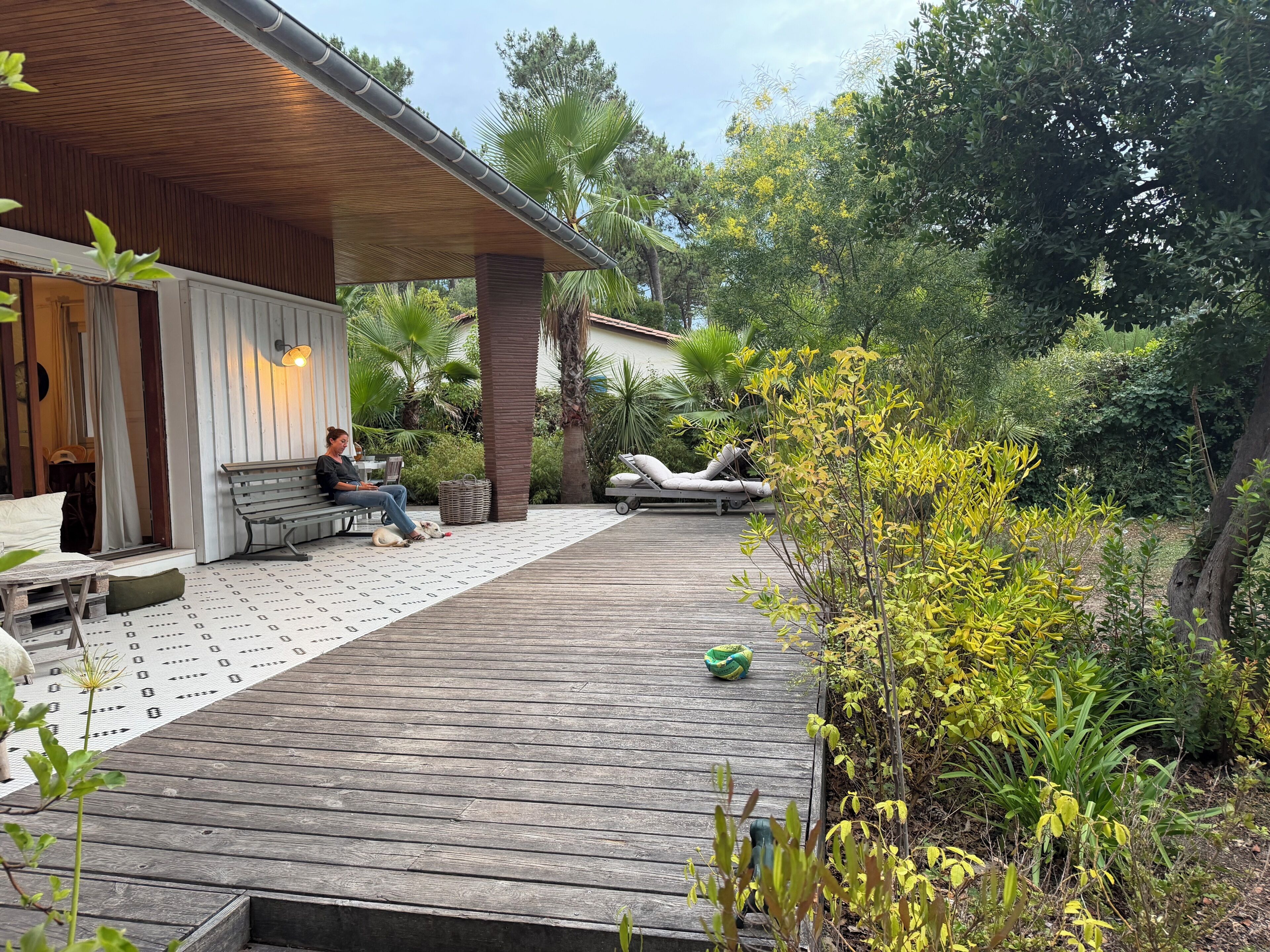 Een houten terras met een bank en een lounge stoel, omringd door groen en bomen.