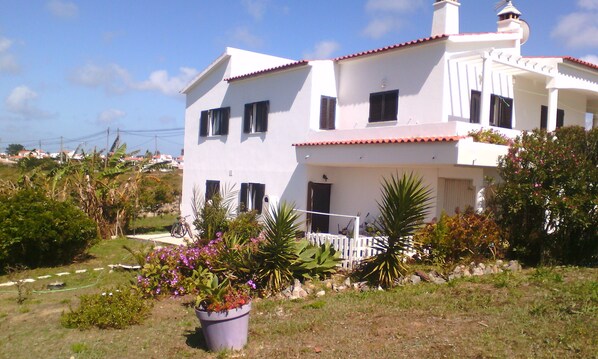 A two-story house with a white exterior and red-tiled roof. There is a balcony on the second floor, and the property is surrounded by greenery and a garden with various plants and flowers.