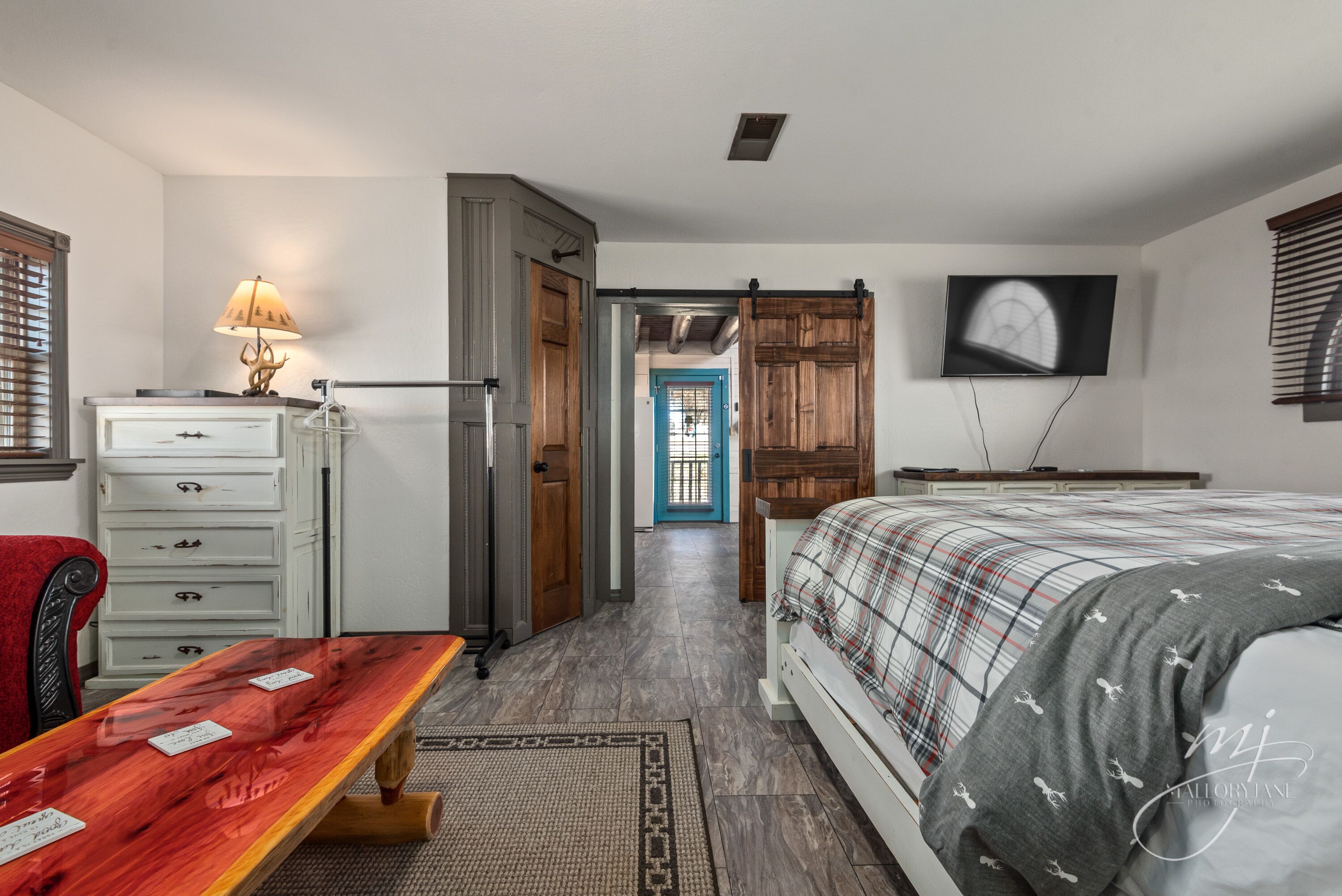 A bedroom with a bed, a desk, a dresser, a TV, and a door leading to another room.