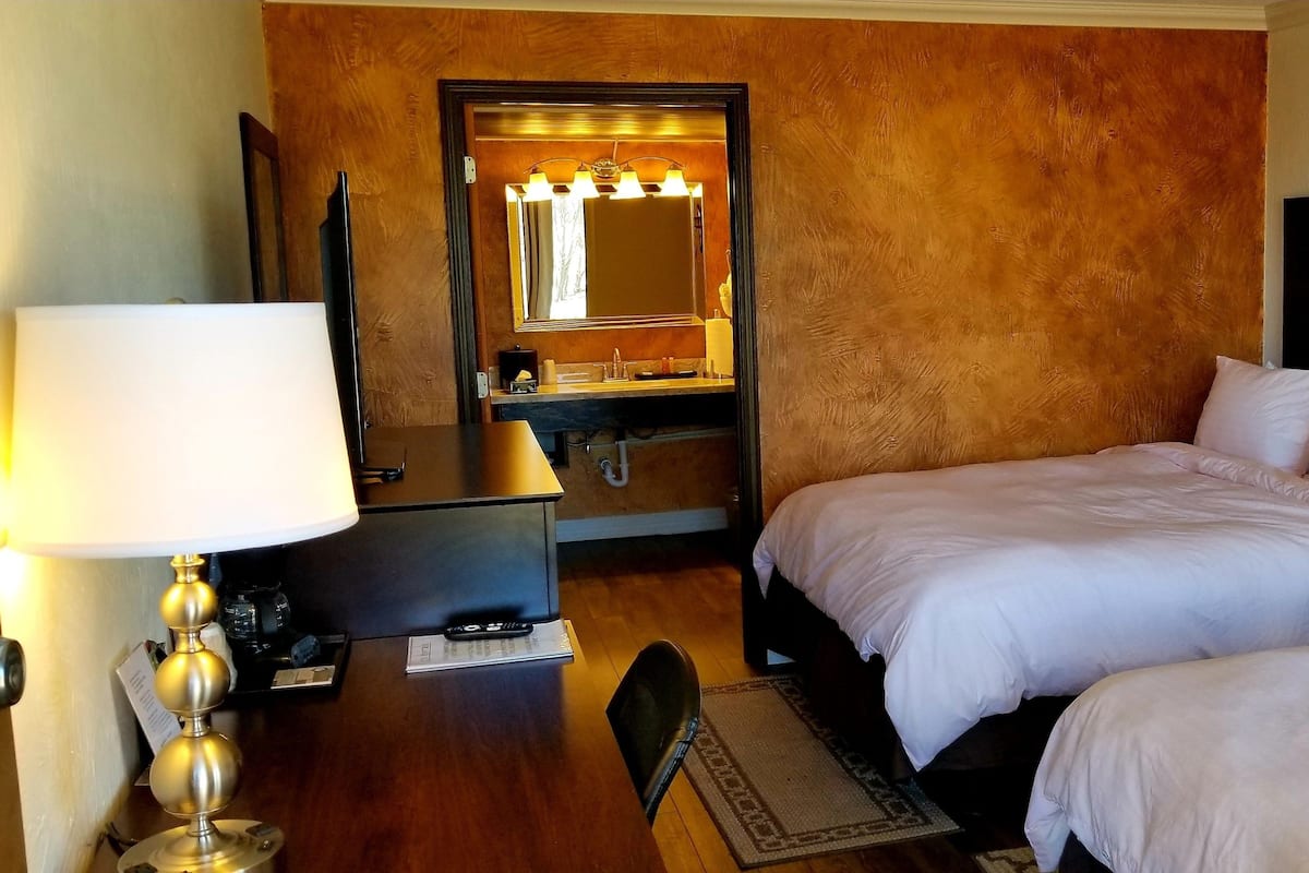 A hotel room with two beds, a desk with a lamp, a vanity with a mirror, and a telephone on the nightstand.
