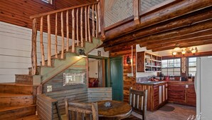 Interior de una cabaña de madera con escalera, comedor, cocina y una chimenea acogedora.