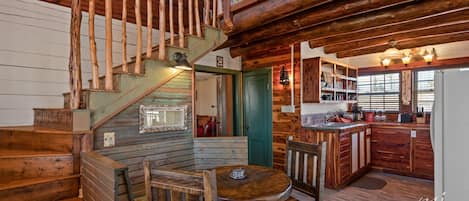 A wooden cabin interior with a staircase, dining area, kitchen, and a cozy fireplace.