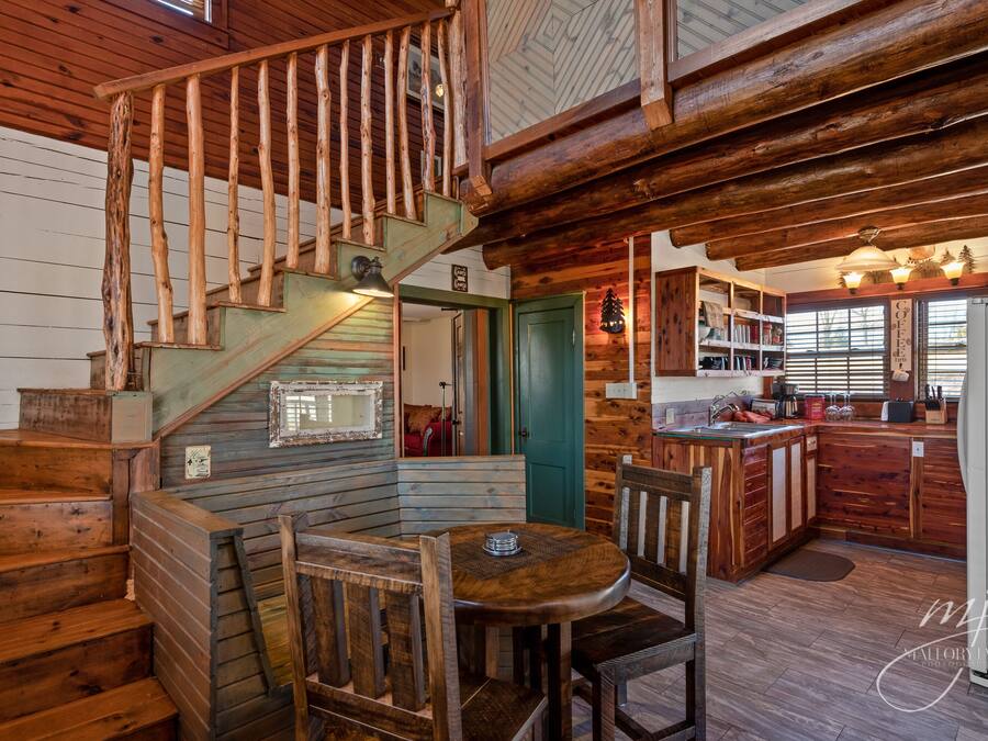 A wooden cabin interior with a staircase, dining area, kitchen, and a cozy fireplace.