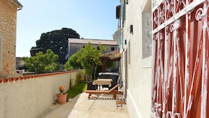 Un patio exterior estrecho con mesa y sillas de madera, una planta en maceta y un portón rojo.