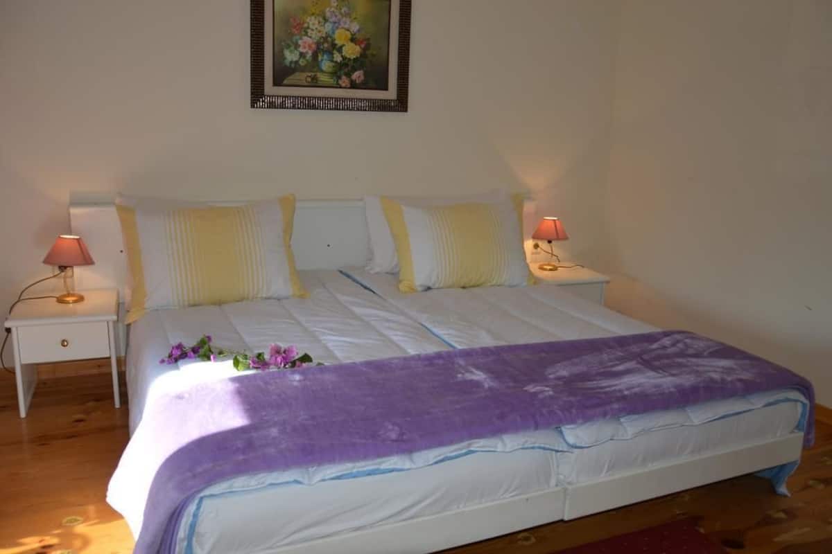 A neatly made bed with a purple blanket, two bedside lamps, and a framed floral painting on the wall.