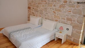 Un dormitorio con pared de piedra, una cama con ropa blanca, una mesita de noche con lámpara y suelo de madera.