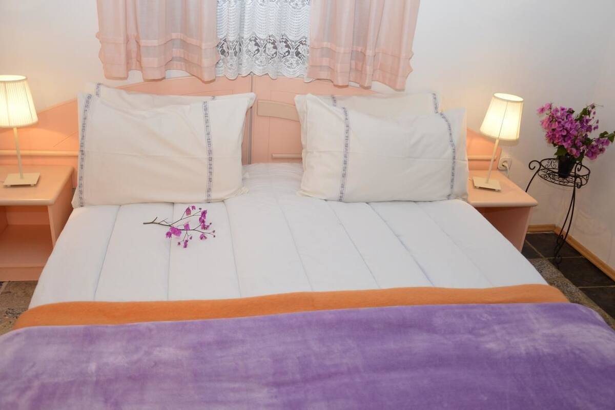 A neatly made bed with white linens and two pillows, a purple throw blanket, and a branch with purple flowers.