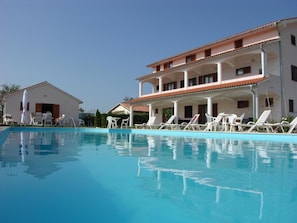 A swimming pool with lounge chairs and a building with a tiled roof and columns.