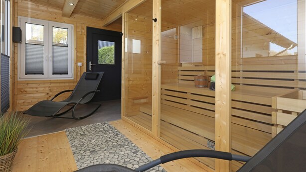 Une cabane en bois dotée d’une porte coulissante en verre donnant sur un espace sauna, d’un fauteuil de relaxation noir et d’une petite table sur laquelle est posée une plante.