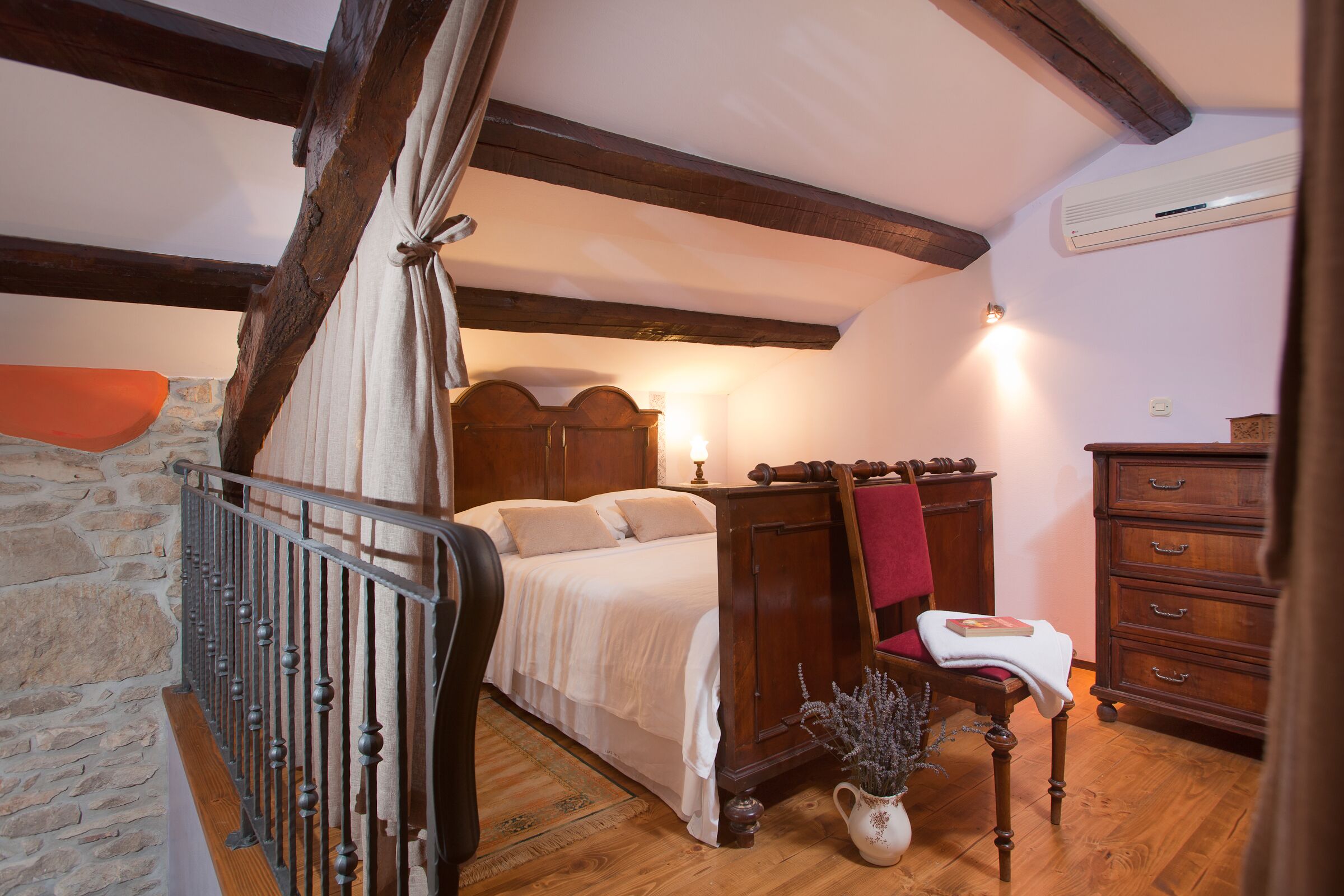 Une chambre à coucher avec un lit en bois, un mur en pierre, une commode en bois et une chaise avec un coussin rouge.