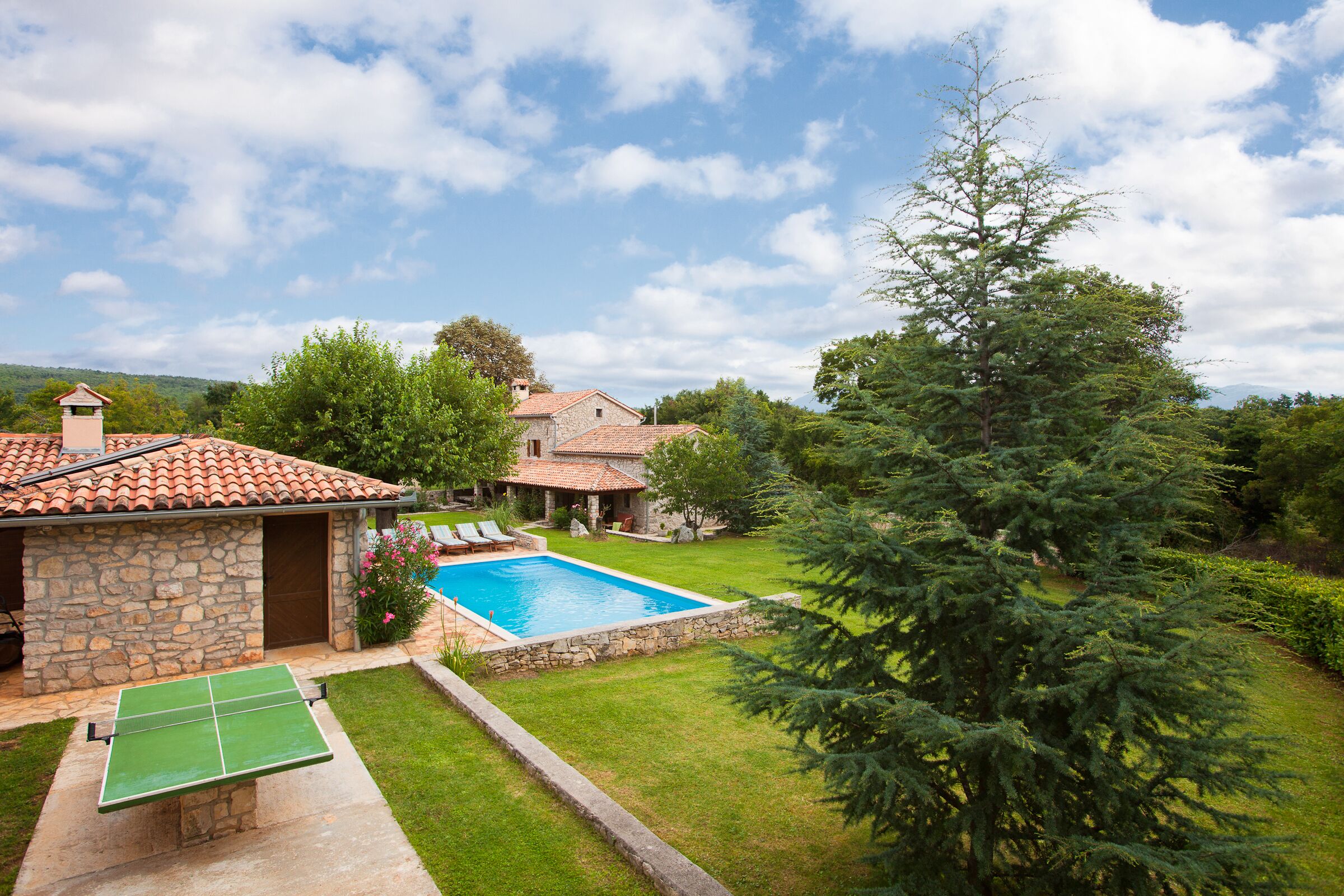 Una casa in pietra con piscina, tavolo da ping pong e un grande albero sempreverde.