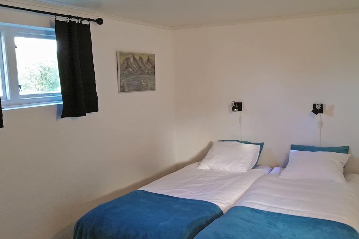 Ein Doppelbett mit weißer und blauer Bettwäsche, ein kleiner Nachttisch mit Lampe, ein Fenster mit schwarzem Vorhang und ein Gemälde an der Wand.
