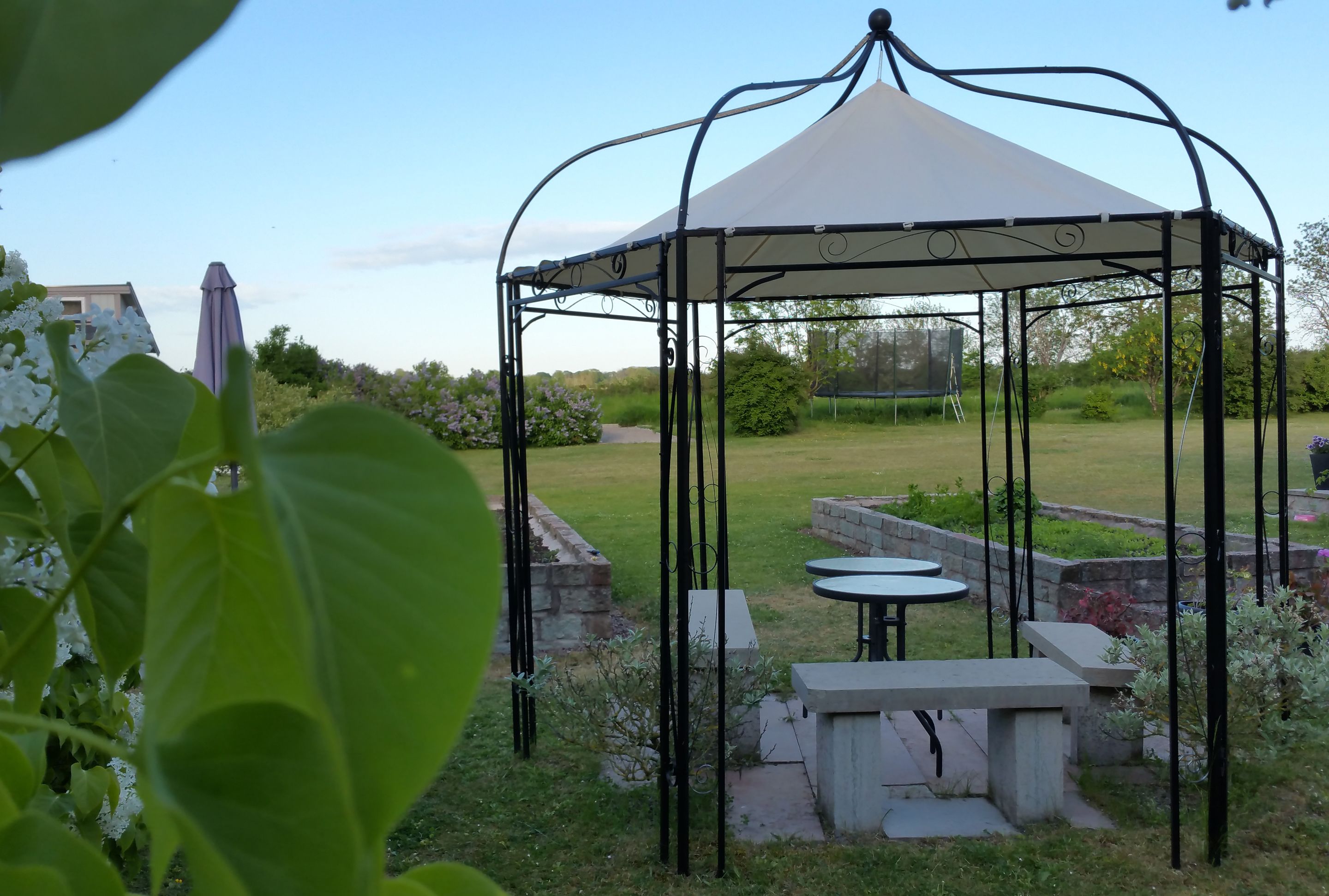 A gazebo with a white canopy and metal frame, surrounded by a garden with a stone bench and a small table.